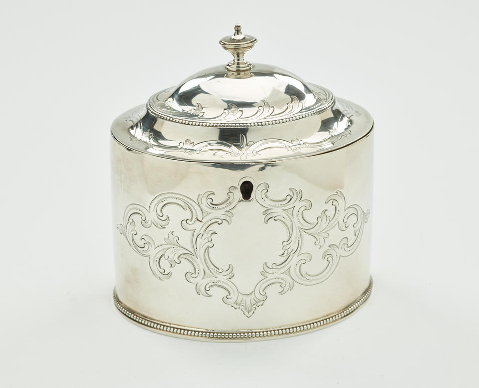 A silver tea caddy with a lid and sculptural finial. There is also swirling engraving along the body and lid.  