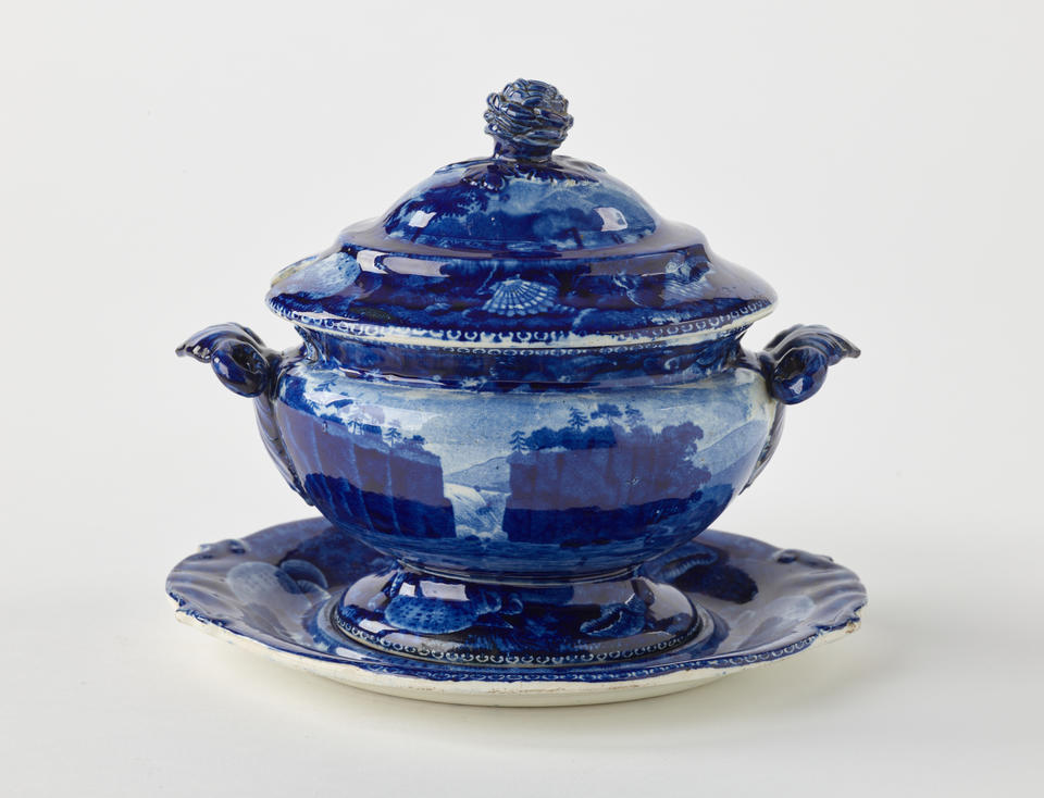 A dark blue decorative sauce boat, two symmetrical sculptural handles that depict a bird’s head, foot, and lid with rounded finial 