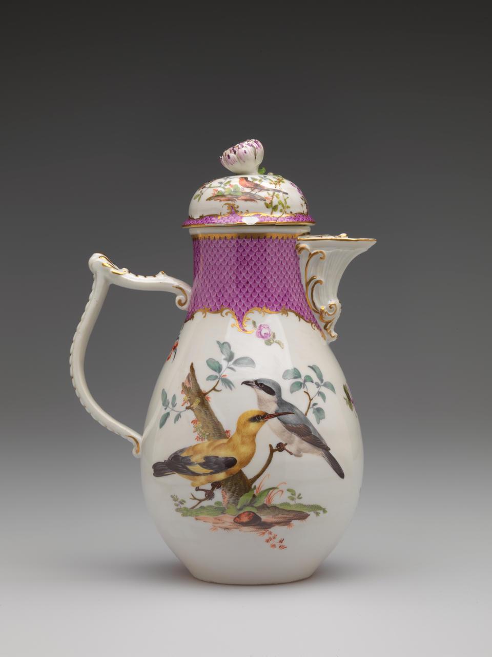 A white coffee pot with a handle and spout, gilded, pink, and floral decorations. There are two birds, blue and white, the other yellow and black, both positioned on branches.