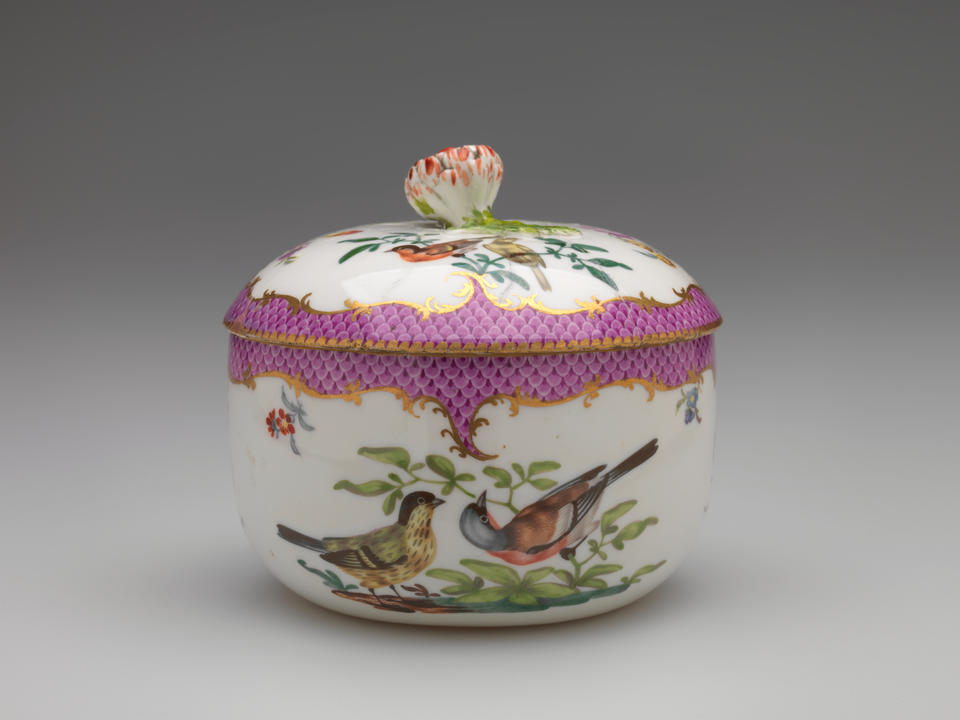 A white vessel with a lid, with  gilded and pink decoration, floral decorations, and two birds that are white, black, red and blue, both are on a branch.