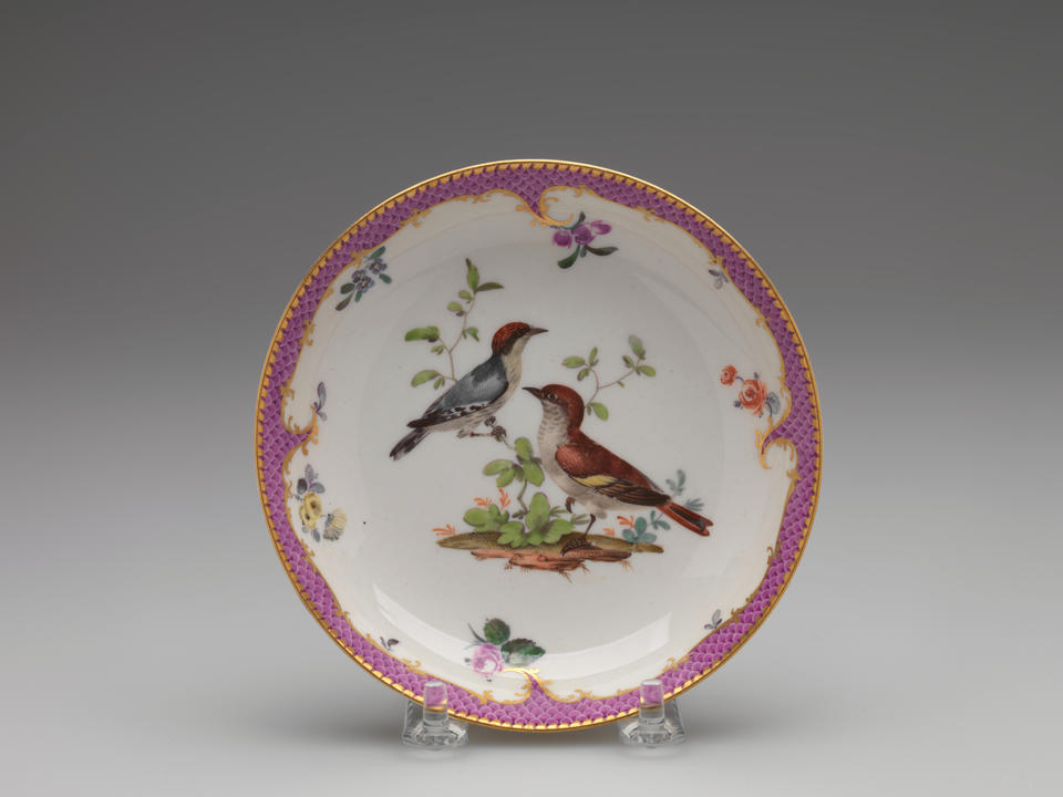 A white saucer with gilded, pink, floral decorations, and two birds; one bird is blue with a red head positioned on a branch, the other is standing on the ground.