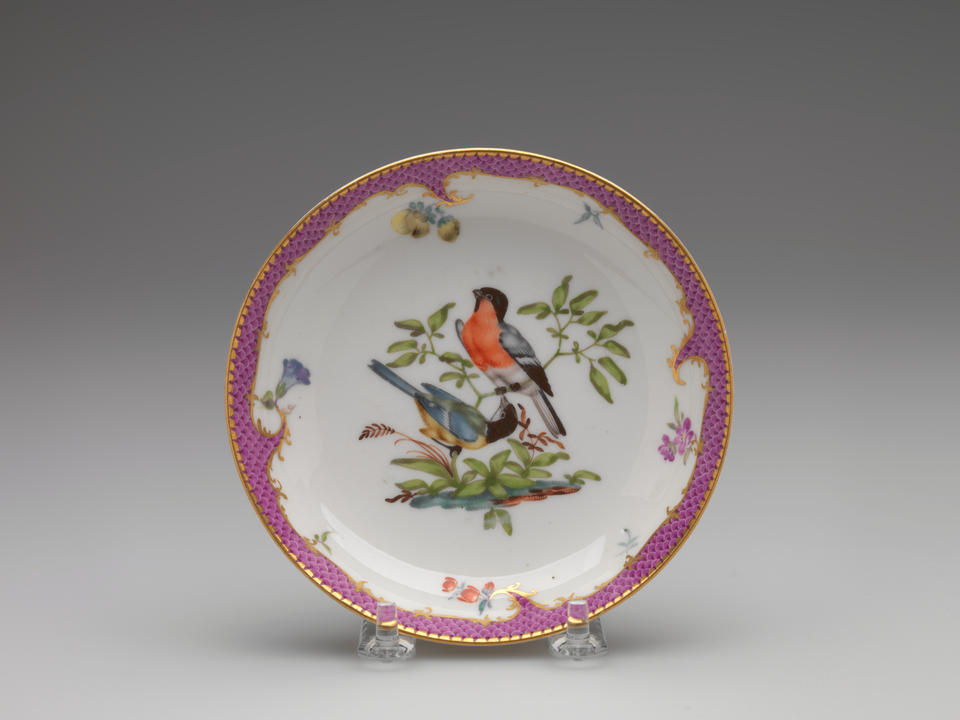 A white ceramic plate with a gilded and pink edge, floral patterns, with two birds surrounded by floral elements, positioned on branches, are blue, white, black, red, and yellow.
