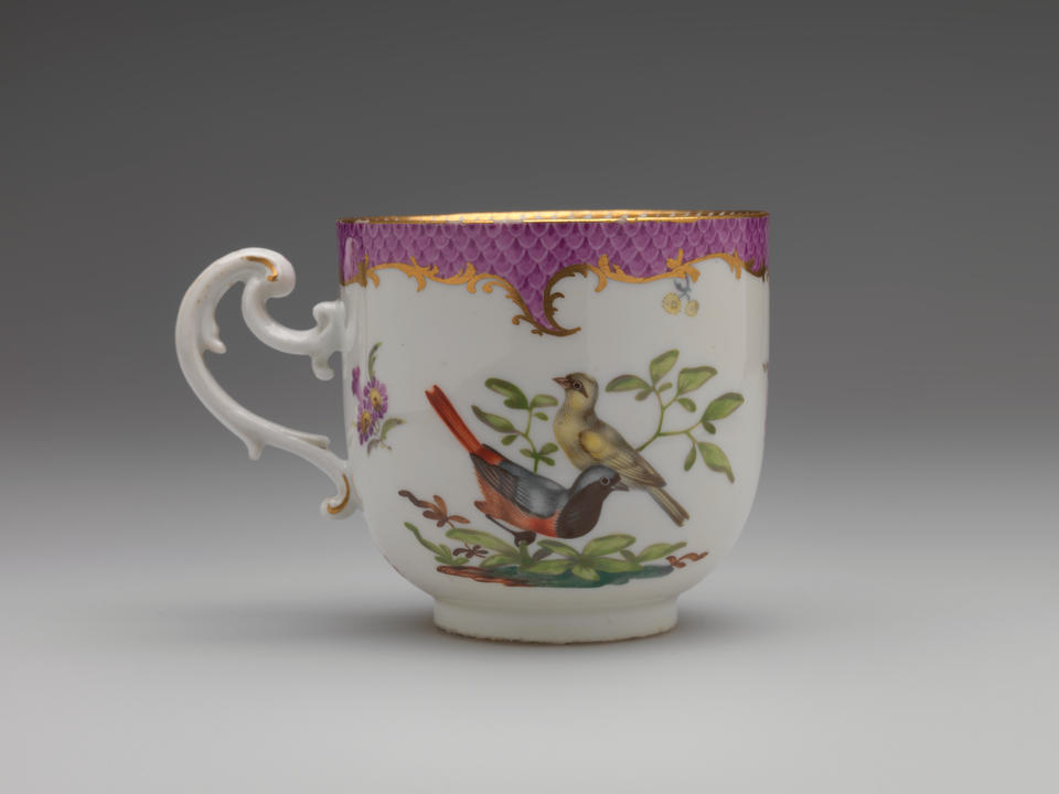 A white teacup with a handle that has swirls, gilding, pink, and floral decorations. There are two birds, one large and yellow, the other is white, black, blue and orange.