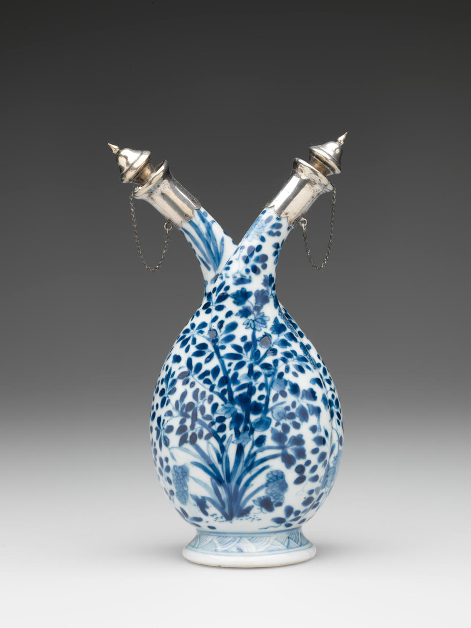 White and blue vessel with a flared foot, rounded body, and two necks in opposite directions. Necks are closed with silver caps attached to the vessel with delicate chains. 