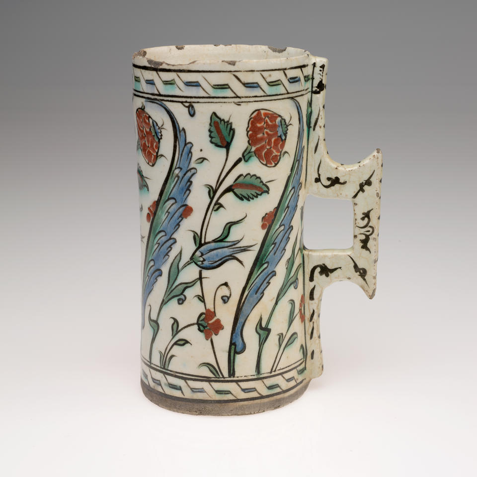 A rounded cream colored vessel with a square handle, decorated with geometric and flower designs in blue, green, and red. 