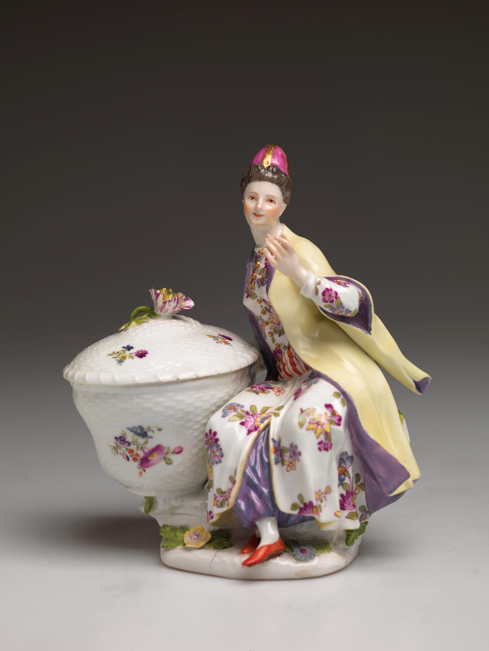 A sculptural figure sitting next to a large basket, the basket and figures clothing has floral decorations. The figure is wearing a hat, cream and purple clothing, and red shoes. 