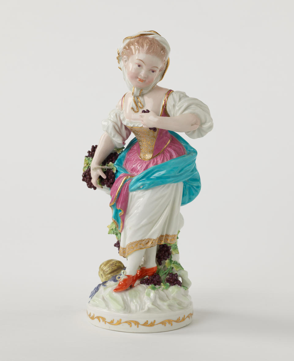 A sculpted porcelain light haired figure with grapes dressed in historical clothing that is gold, white, blue, pink, and red. 
