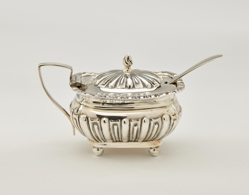  A silver, glass, and gilded mustard pot with a protruding spoon, the lid is hinged to the angular handle. The decorations are raised. 