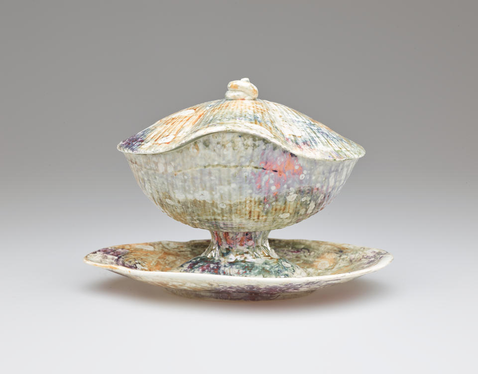 A lidded ceramic vessel on an organically shaped tray. A small, swirled finial is on the lid. The body of the vessel has ridges covered with a mottled, multi-colored glaze.