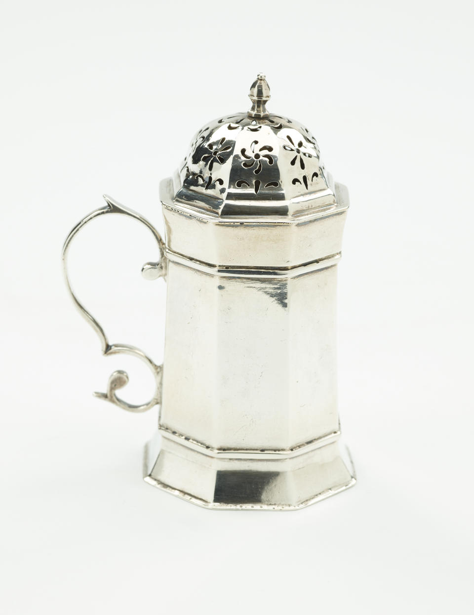 A silver caster that has an octagonal body, handle, and lid that has floral perforations.