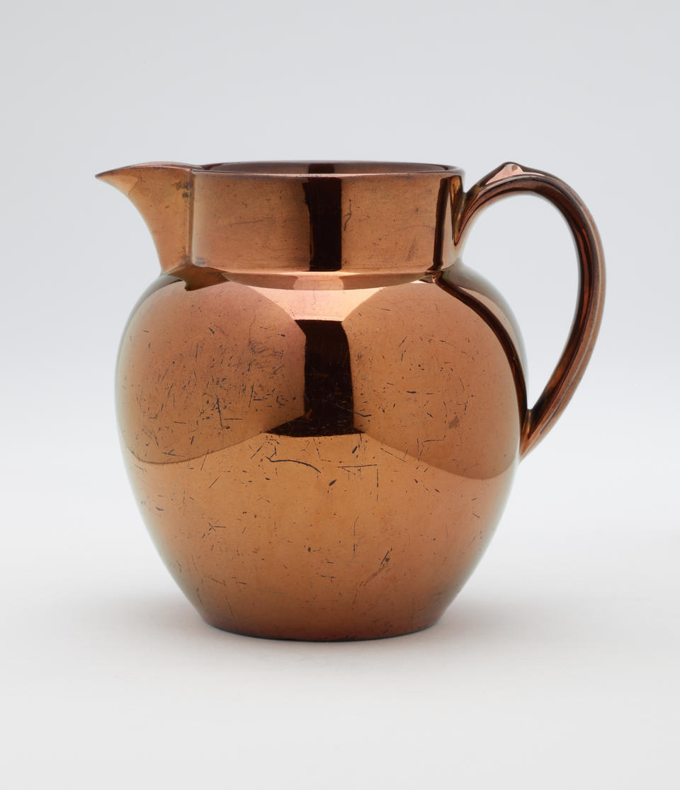 A ceramic jug with a small handle. The glaze is a brown metallic color.