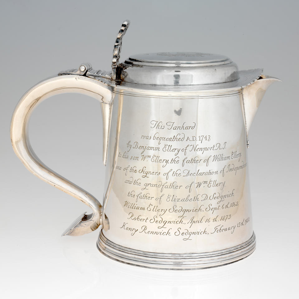 A rounded silver vessel with a curved handle, lid, and spout. Side of the vessel is covered with engraved writing. 
