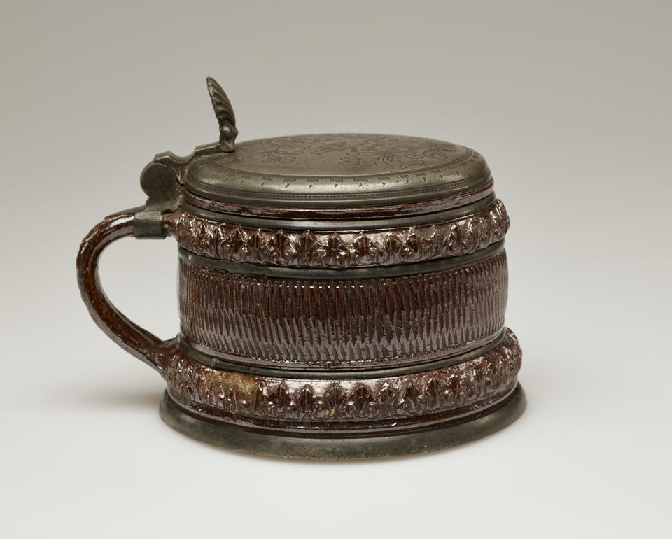 A short metallic glazed stoneware tankard with a delicate handle and hinged lid. There is abstract repeating decoration along bands on the entire vessel.