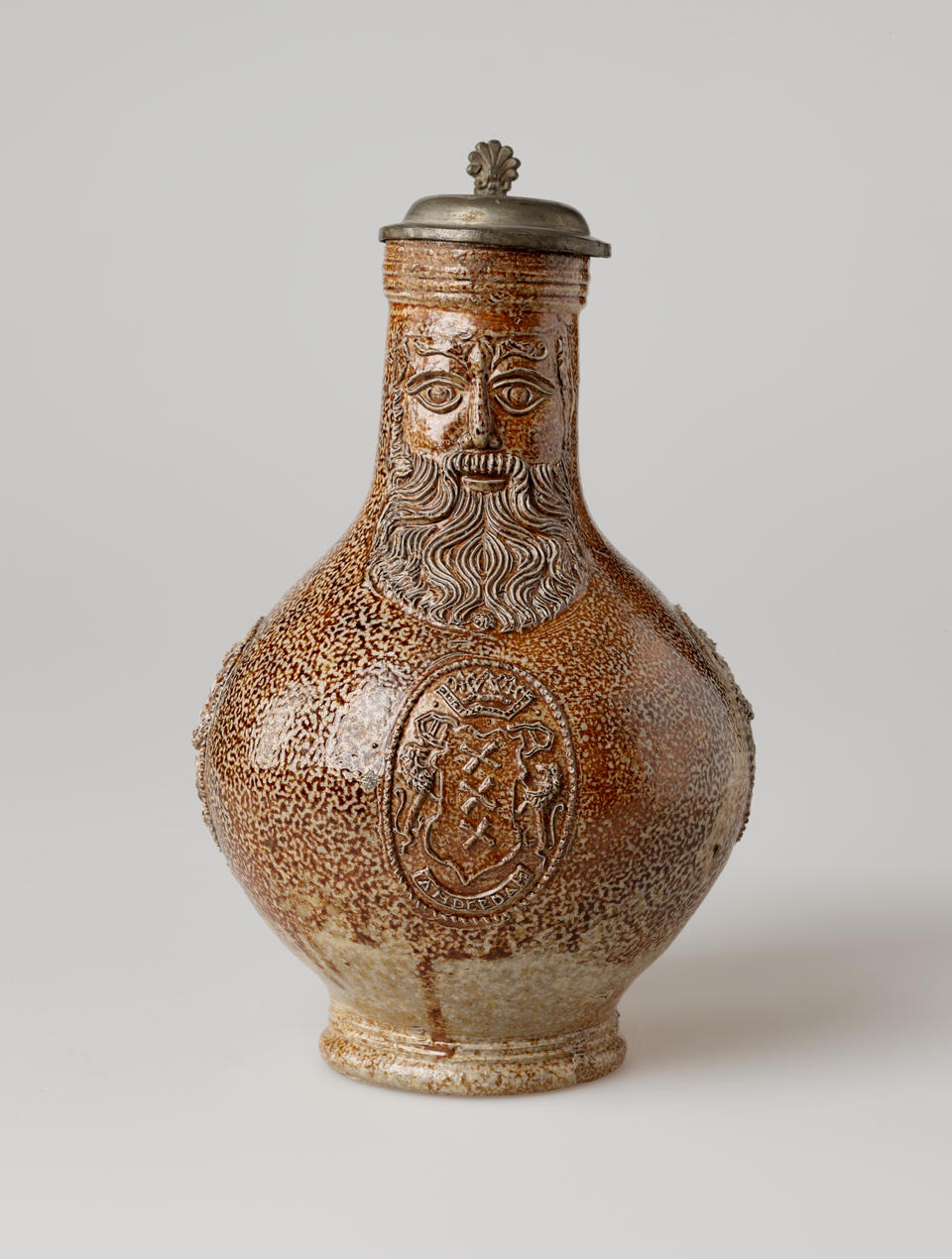 A brown stoneware tankard with a thick handle and metal lid with delicate decorative elements depicting a face and heraldry symbolism.