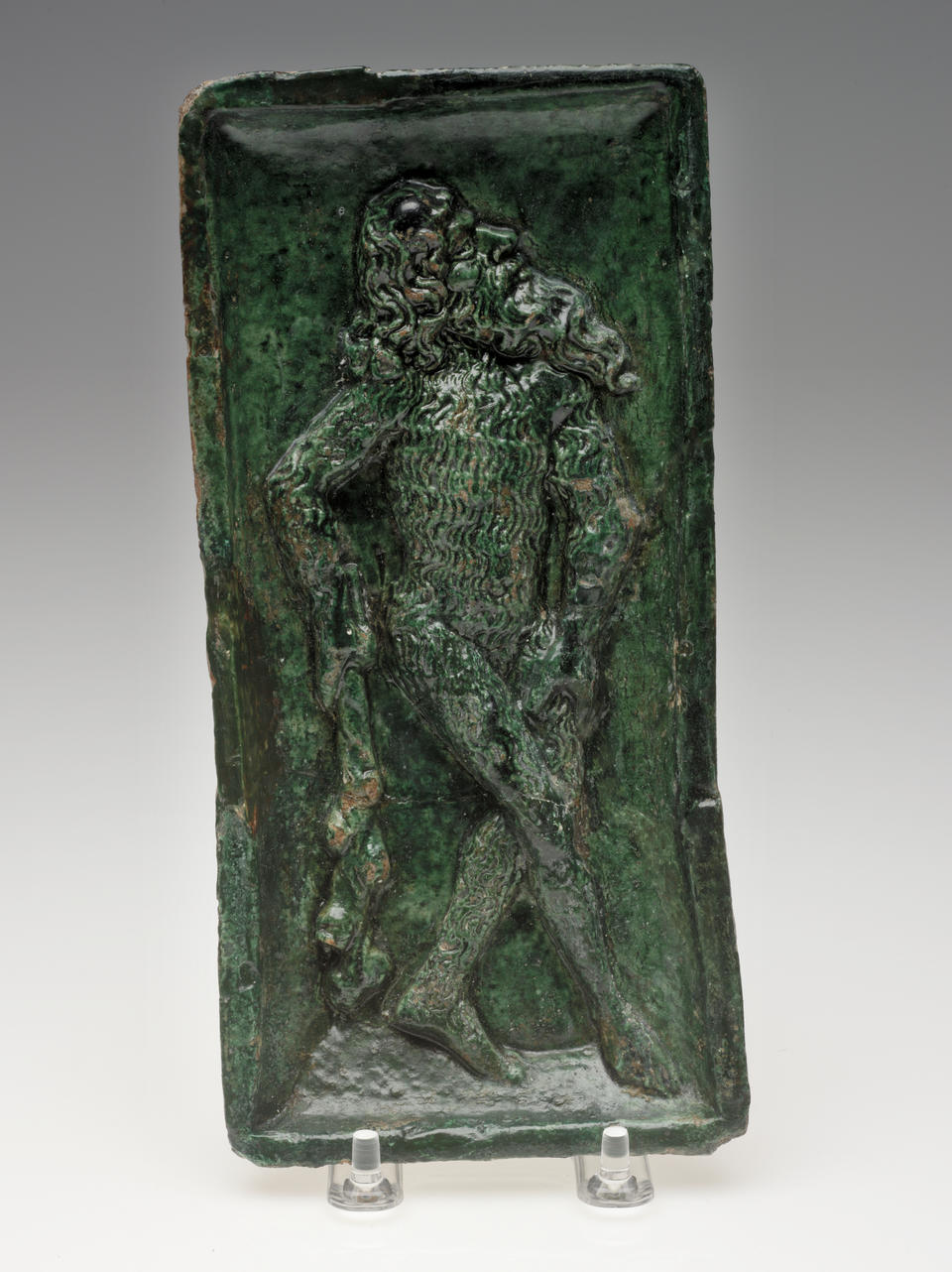 A green sculpted tile of a figure with long hair and beard, holding a stick-like object.