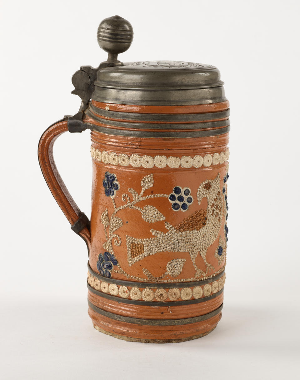 An orange earthenware stein with a metal lid that is hinged to the handle.  The white and blue decorations consist of dots and are primarily depicting floral imagery.