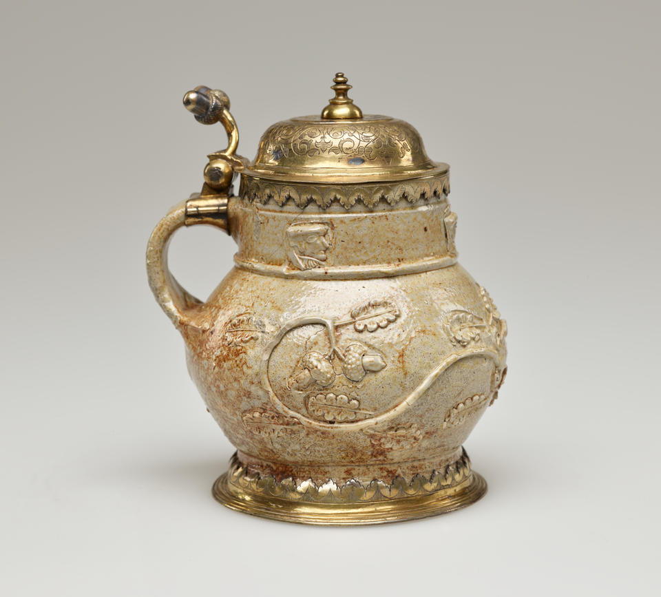 An unglazed stoneware mug with tan to copper colors and floral decoration. The foot goes into a gilded metal decoration, similarly to the handle where the hinged lid is attached.