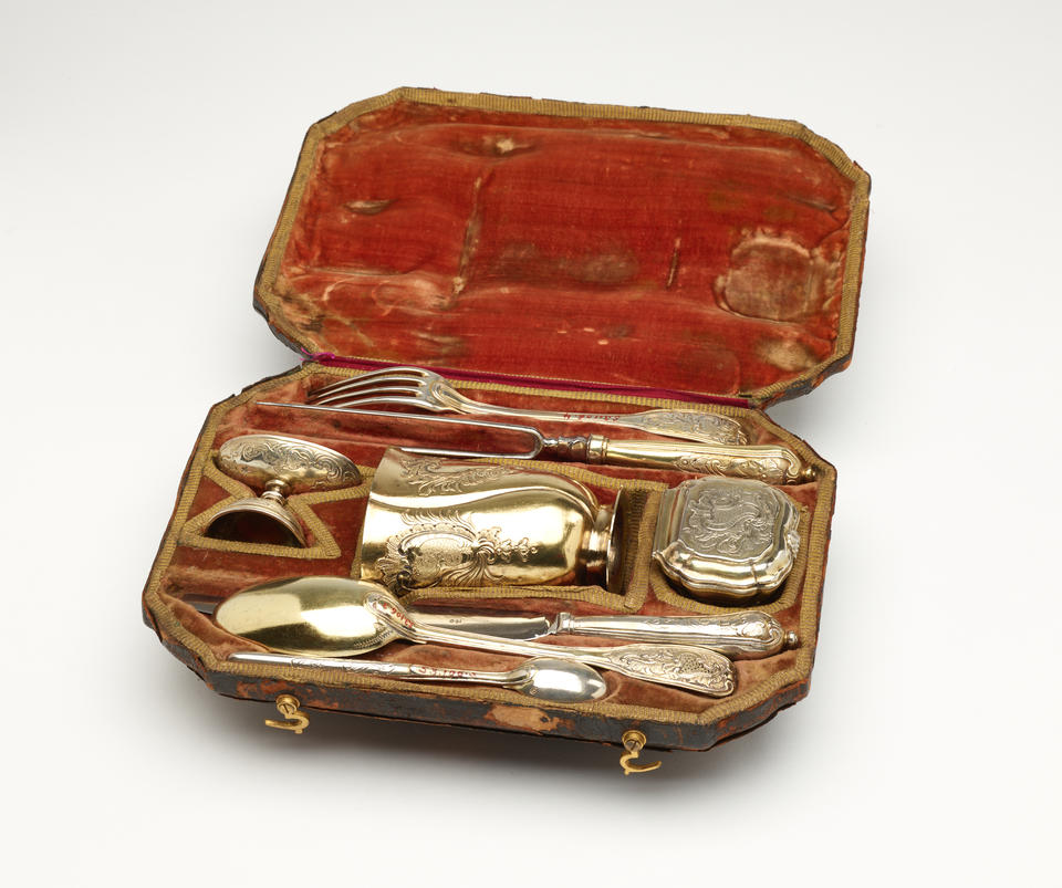 A traveling set with silverware. The traveling case is an organic colored fabric. There are multiple indents holding the silver, steel, and gilded cutlery and vessels.