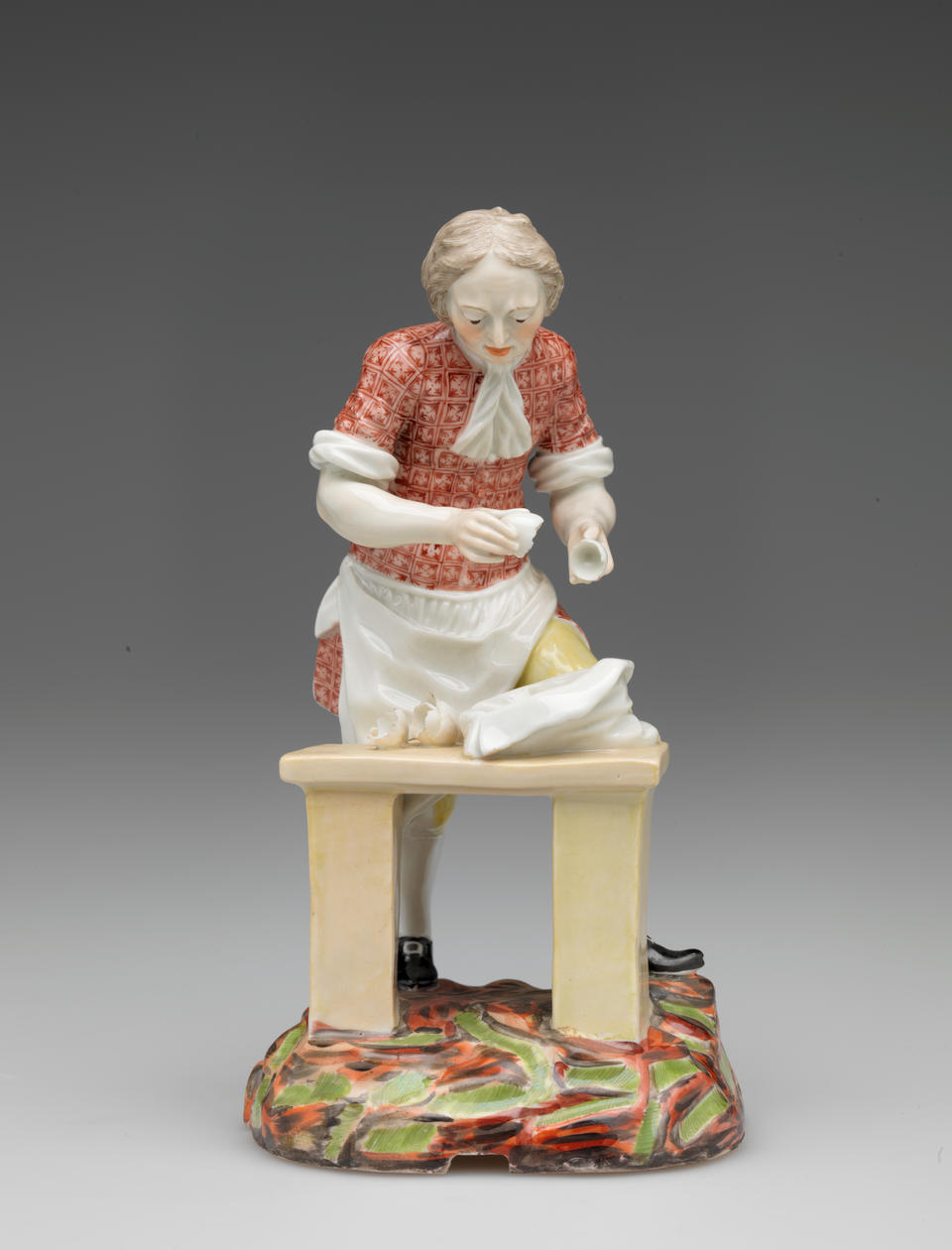 A sculpture of a figure in historical clothing and a table, a sack, and broken egg shells. The ground is brightly colored red, brown, and green. 