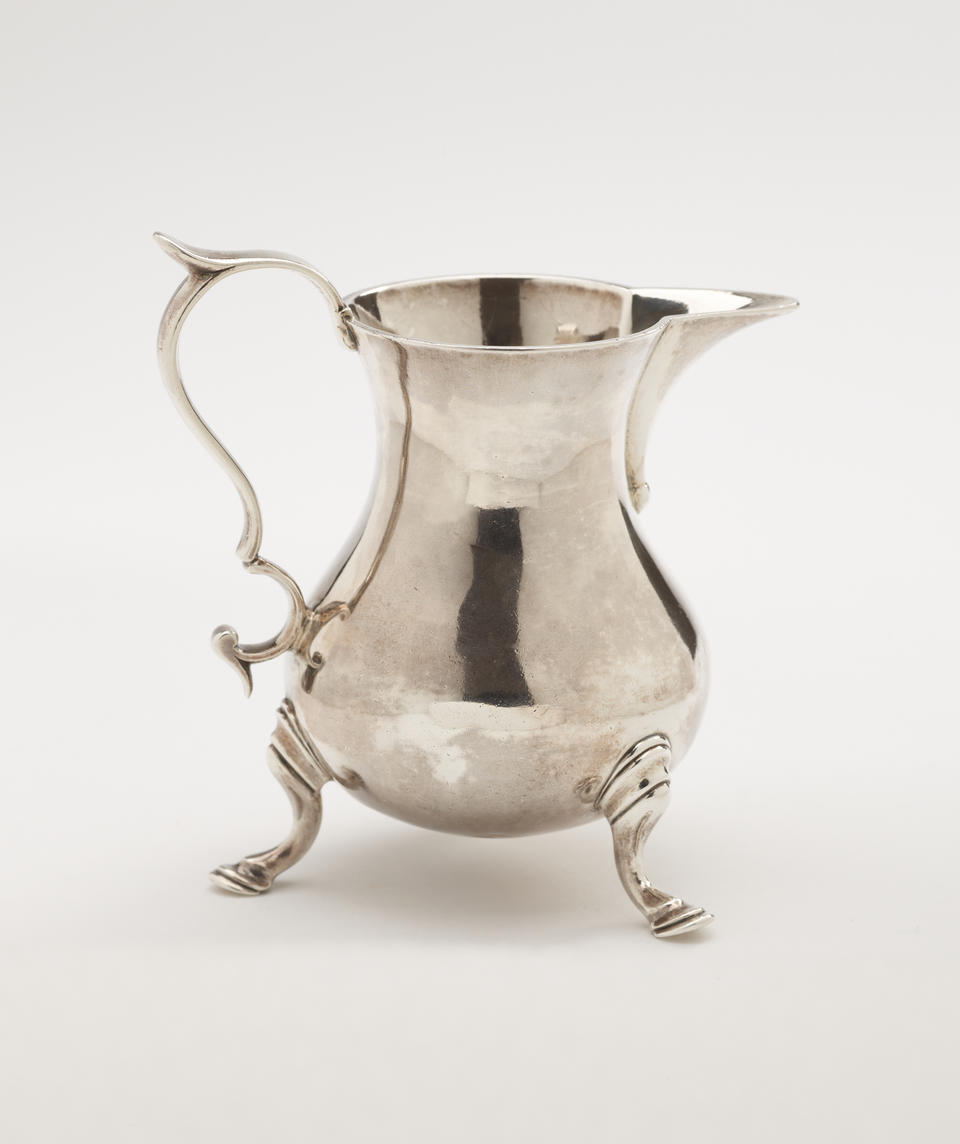 A silver creamer with a sculptural handle and three protruding feet. 