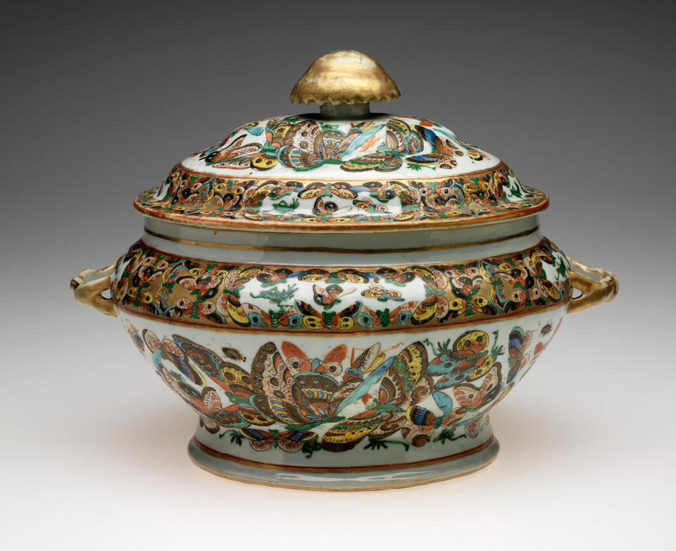 An oval lidded vessel with a large gold finial. The body of the vessel is decorated with multicolored butterflies on a white ground. 