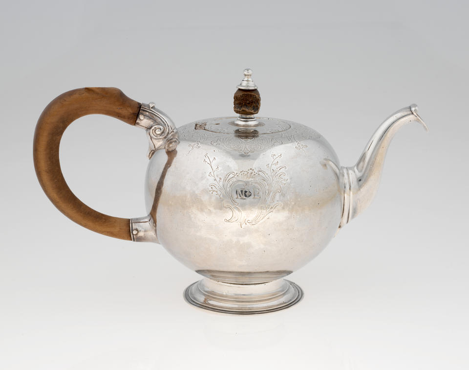 A bulbous silver teapot with a long curved spout, and foot, wooden finial and handle connected with decorative silver pieces. There are delicate engraved marks on the body and lid.