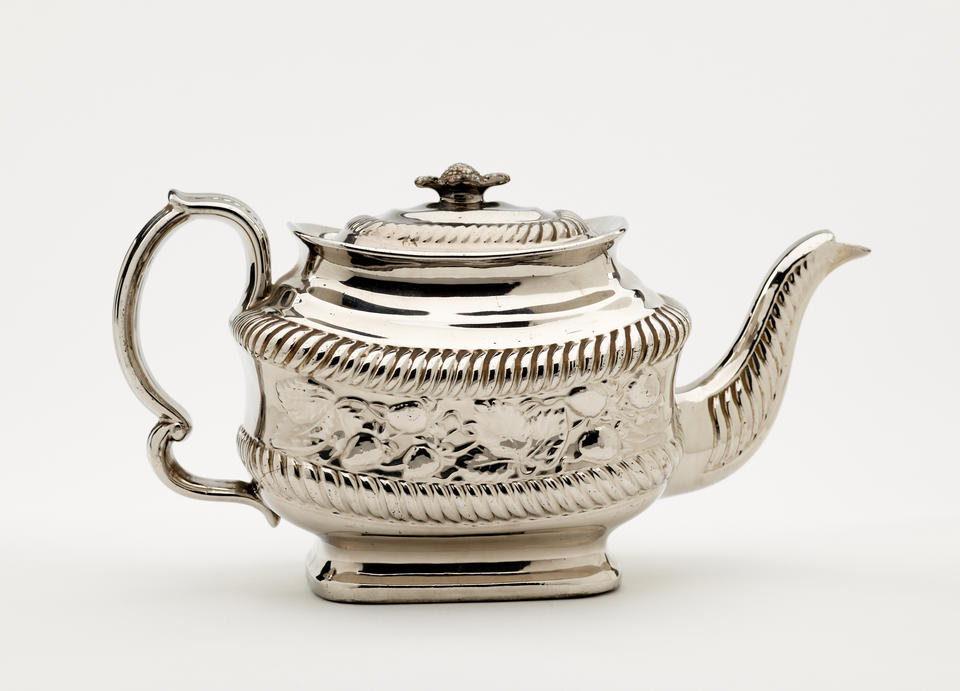 A silver luster teapot with a decorative handle, a rounded square body and spout with sculptural decorations.