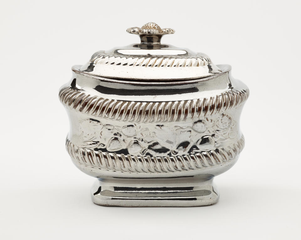A silver luster sugar bowl with a decorative lid and a rounded square body with sculptural decorations.