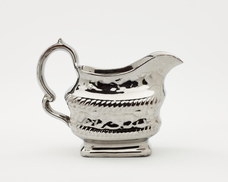 A silver luster creamer with a decorative handle, a rounded square body with sculptural decorations.