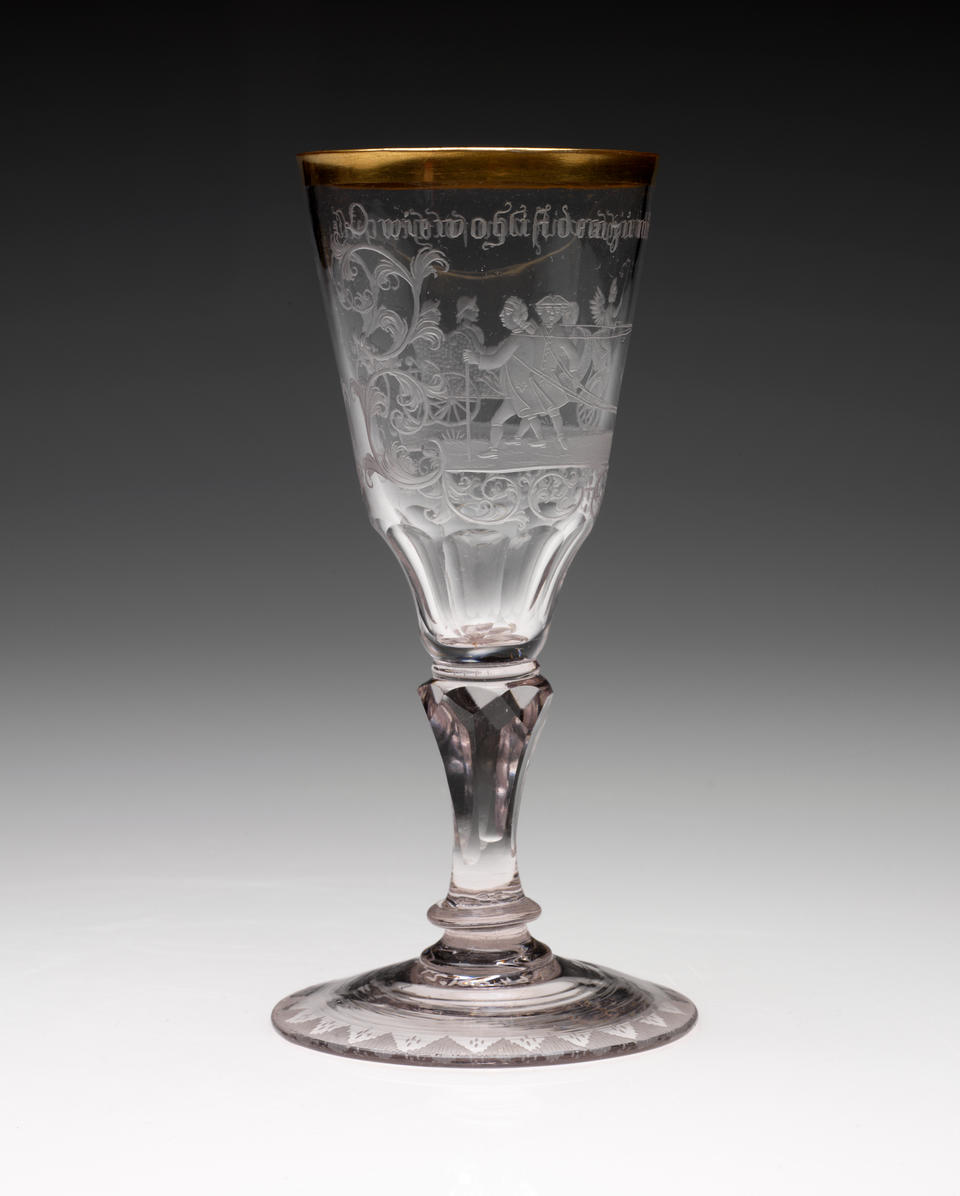 A clear stemmed glass with a rounded foot and thick gold rim. An image of figures walking in a group with words above them are etched in the sides. 