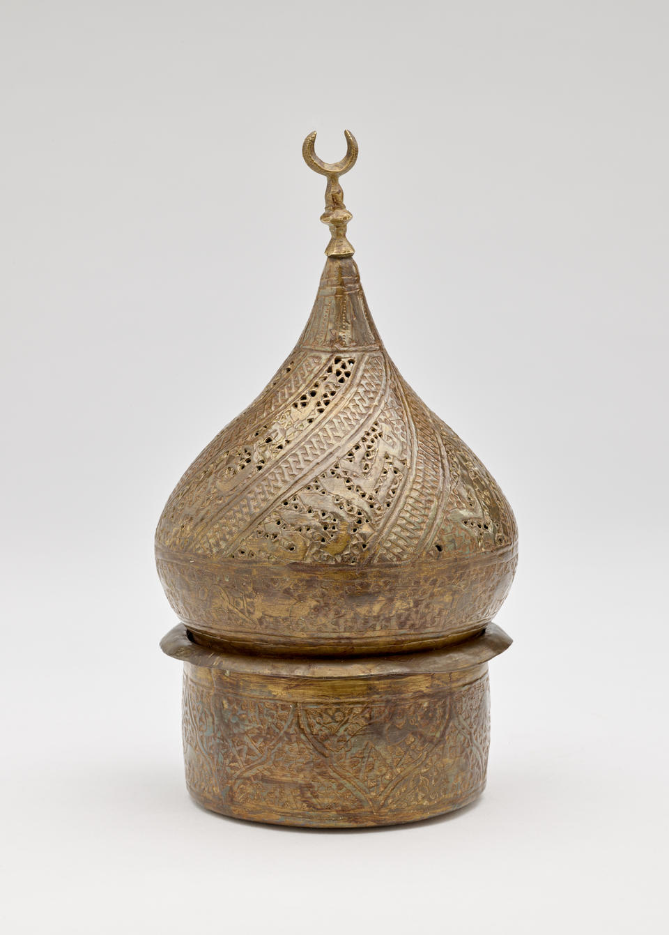 Bronze-colored sculptural object with incised design. Top half is teardrop shaped with a crescent moon shape on top. Cylindrical vessel on bottom. 