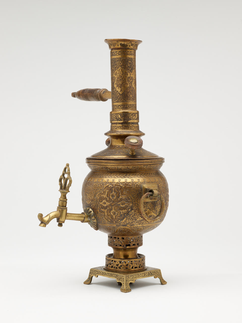 A brass and wood tea instrument that has a decorative foot, lid, various handles and rings, and spout. The entire vessel has highly decorative engravings. 