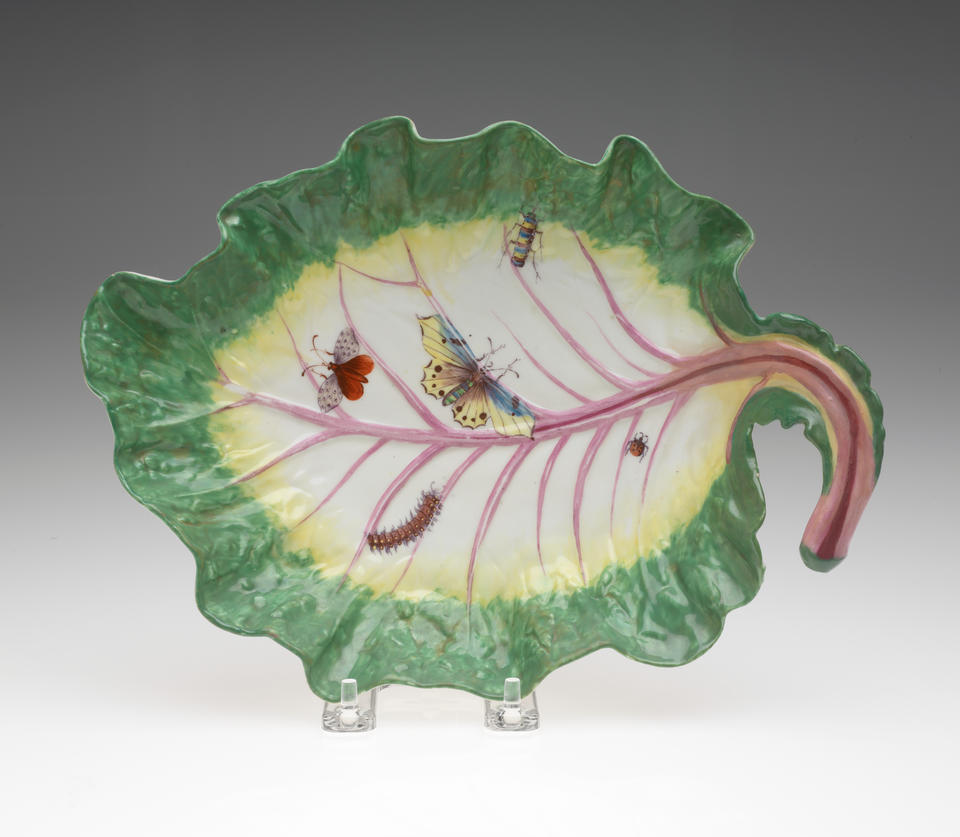 A leaf shaped porcelain dish, green on the edge to yellow then white, the stem is pink, with five insects depicted: a lady bug, two butterflies, a caterpillar, and beetle.