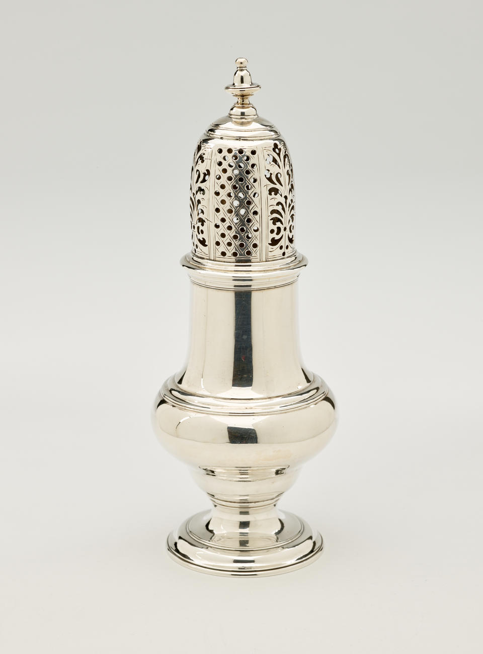 A silver muffineer with a foot, rounded body that goes to a cylindrical top half, and the lid is perforated with small holes and floral cutouts. 