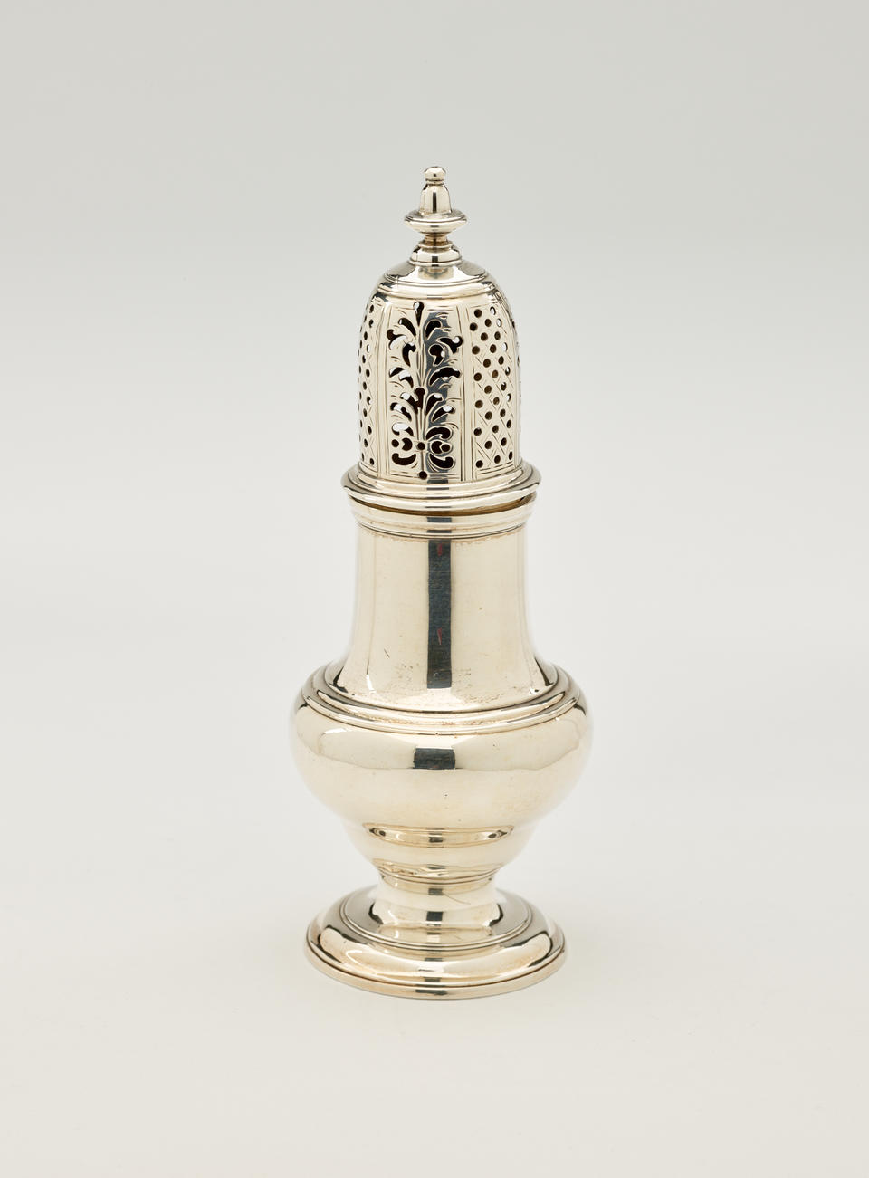 A silver muffineer with a foot, rounded body that goes to a cylindrical top half, and the lid is perforated with small holes and floral cutouts.