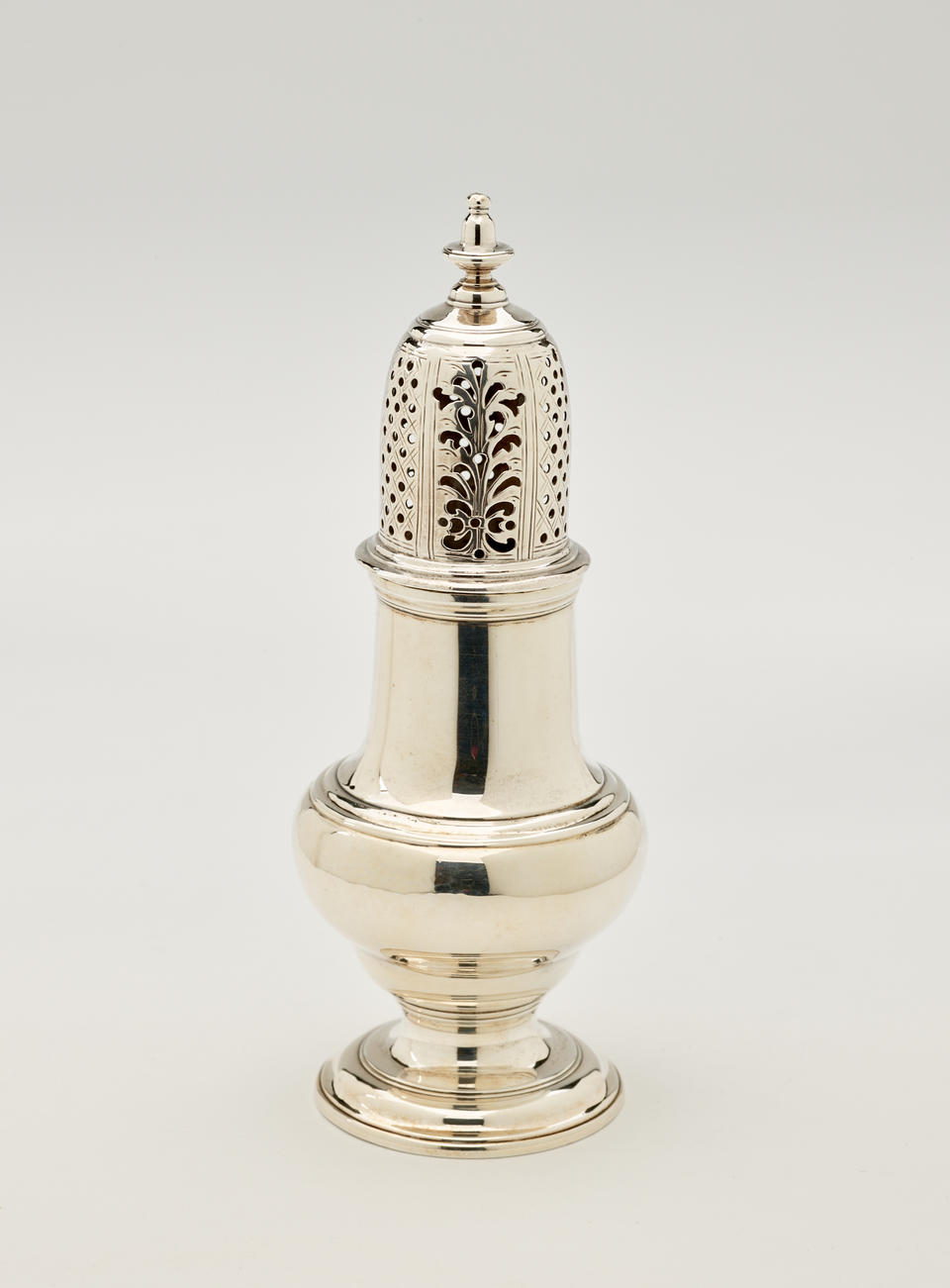 A silver muffineer with a foot, rounded body that goes to a cylindrical top half, and the lid is perforated with small holes and floral cutouts.