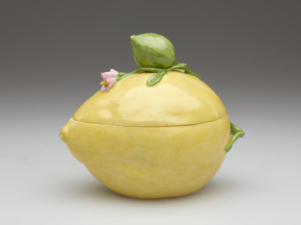 A porcelain lemon shaped box. It is light yellow and has sculpted floral elements such as a pink flower and green bud.  