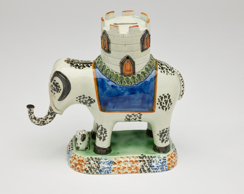 A prattware money box in the shape of an elephant with a castle on its back. The decorations are white, blue, green, and yellow.