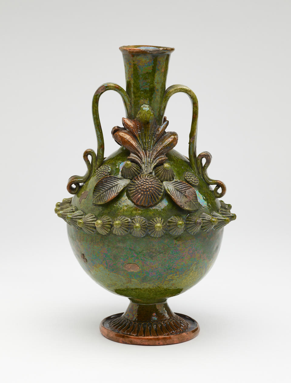 A dark green jug with sculpted decorations, symmetrical decorative handles, and a foot. 
