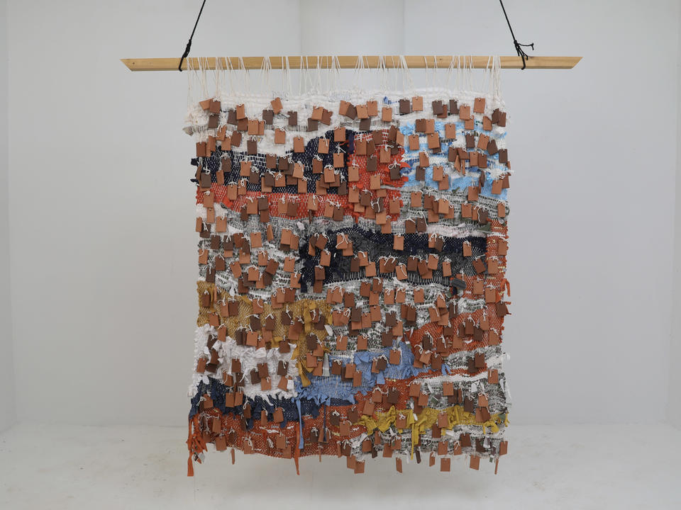 A large, colorful woven textile made of discarded fabrics and old, childhood clothing covered in terracotta tiles hangs