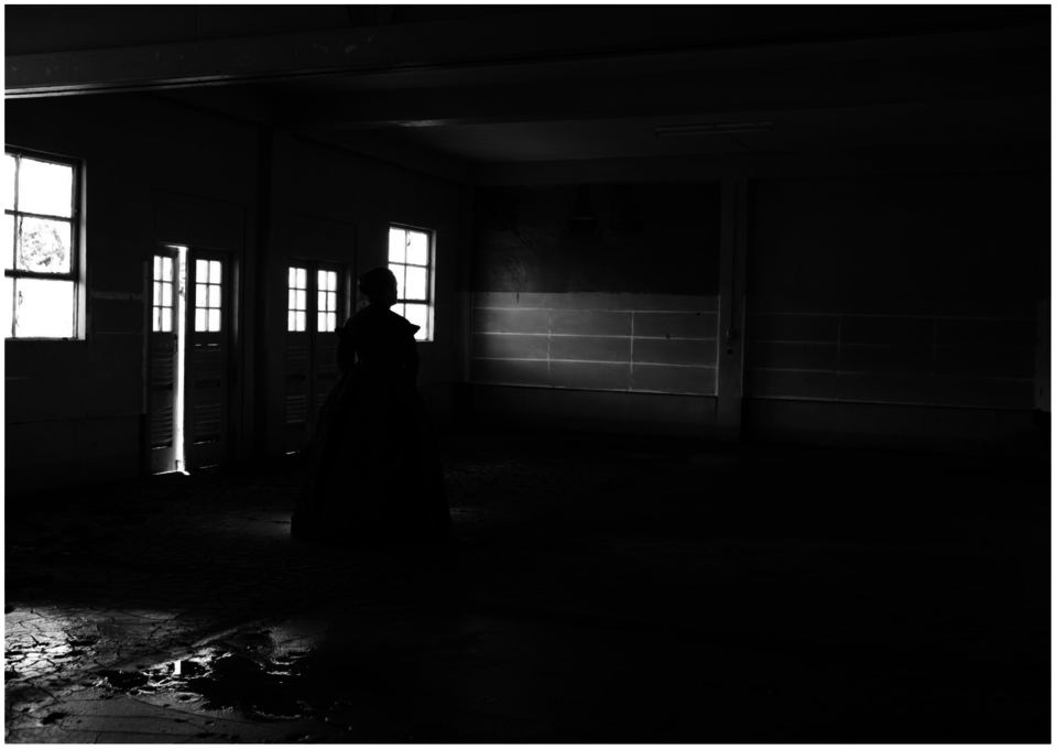 Yuki Kihara's photograph Marist Brothers Old Pupil’s Association House After Cyclone Evan, Mulivai is a black and white indoor scene where a female presenting person in a black Victorian style gown faces an empty wall of an abandoned and poorly lit room.