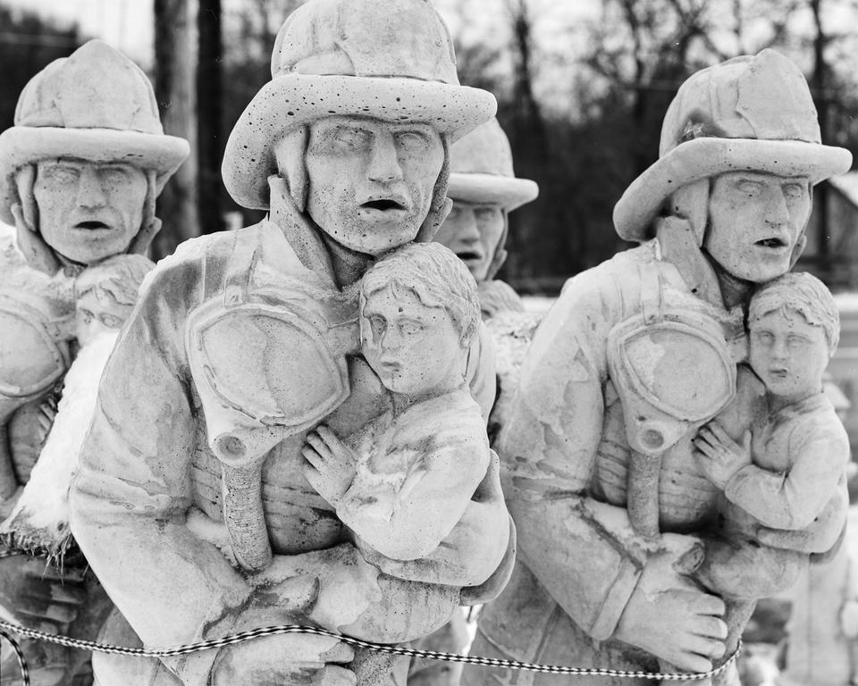 A group of identical white statues of firefighters carrying a child. A rope at the bottom of the frame binds them together.