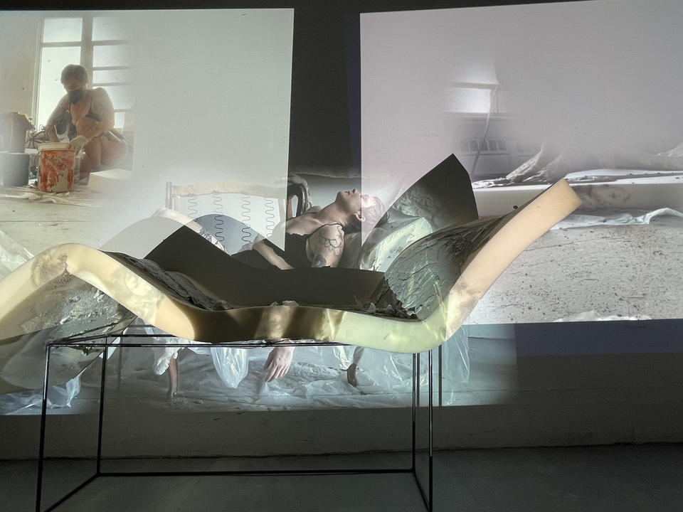 A Triptych of Projections showing the process of pouring concrete on foam, draping the foam across the arms of a chair and then lying over it. These three projections are over a wall and the hardened concrete foam piece. 