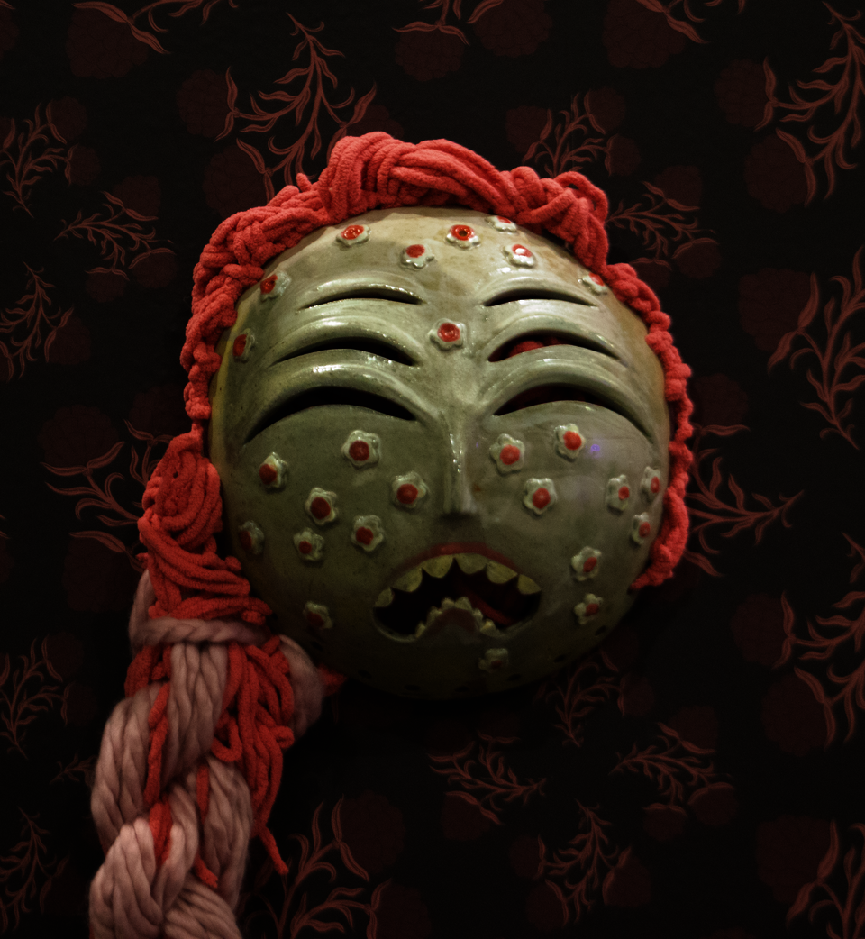 Green ceramic mask with six eyes, wailing mouth, and coral yarn hair.