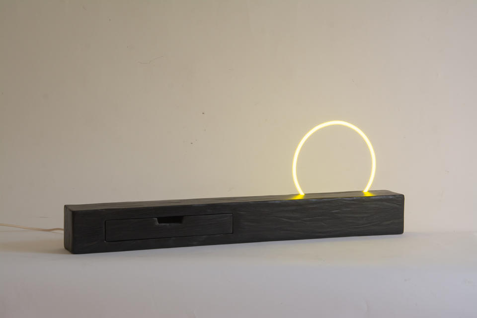 A long rectangular wooden form with an organic carved texture. A perfectly circular yellow neon protrudes from the top of the shelf. 