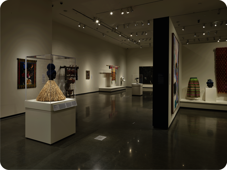 Installation view of an exhibit. A partial wall with a black painted side extends towards the viewer. Visible are colorful textile works, paintings, ceramics, and figurative sculptures. 