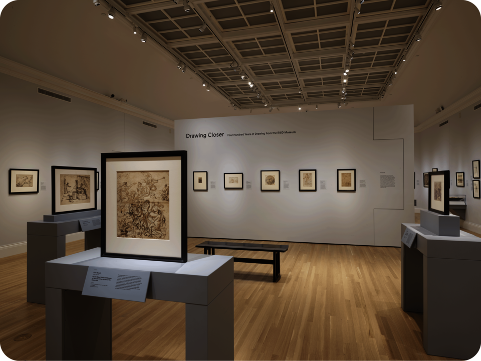 Installation view of an exhibit, with several drawings propped on gray tables, and other drawings hung on the wall. A central divider reads “Drawing Closer”. In front of it is a bench. 
