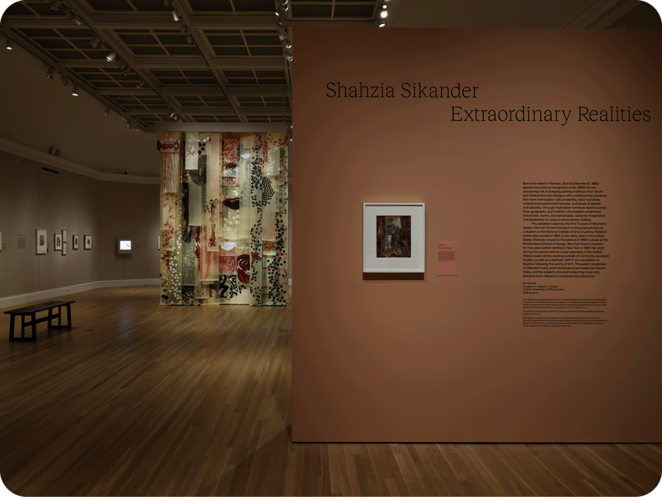 Exhibition documentation. A partial wall in the foreground features artwork and exhibition information, reading “Shahzia Sikander Extraordinary Realities”. Behind it are more artwork on walls, and a larger work suspended from the ceiling. 