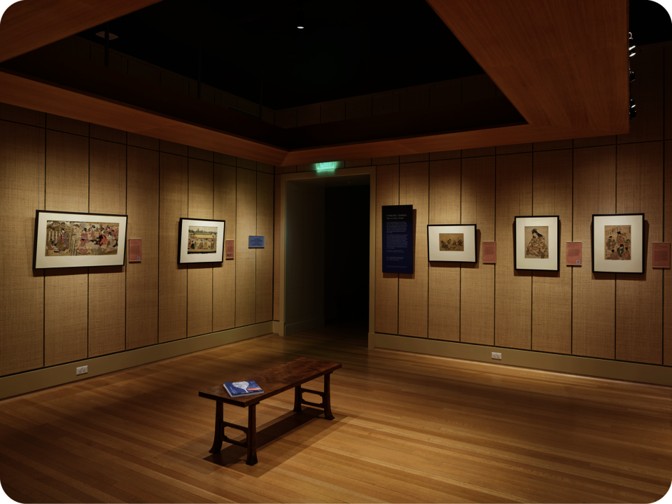 Exhibit documentation. Two framed prints hang on one wall, with three other prints and exhibition text on another wall, separated by a doorway. A wooden bench occupies the space’s center. 