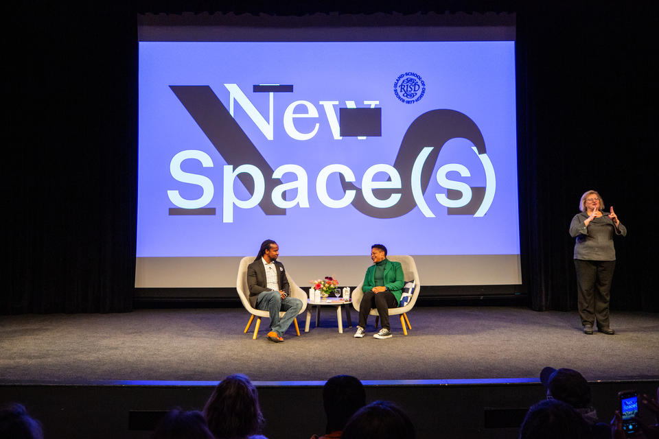 A Black man and woman sit on a stage, conversing. The New Space(s) logo is projected behind them, and an ASL interpreter stands to the side.